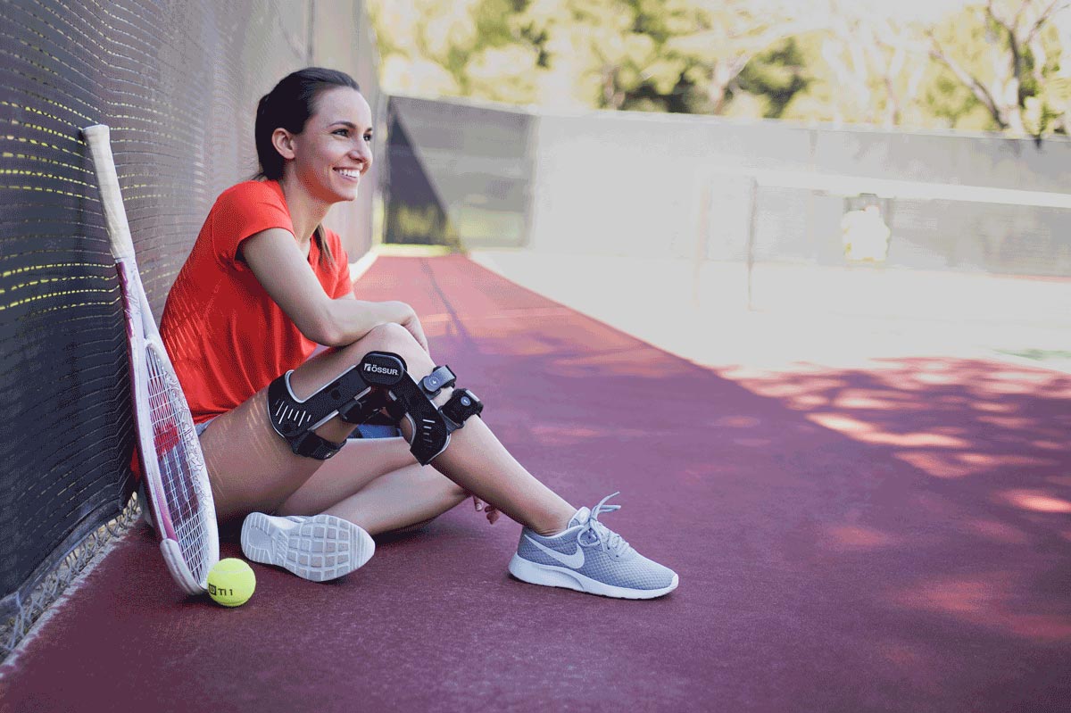 Woman smiling while sitting on a tennis court, wearing a knee brace and athletic shoes, with a tennis racket and ball nearby, illustrating support for sports injuries and mobility.