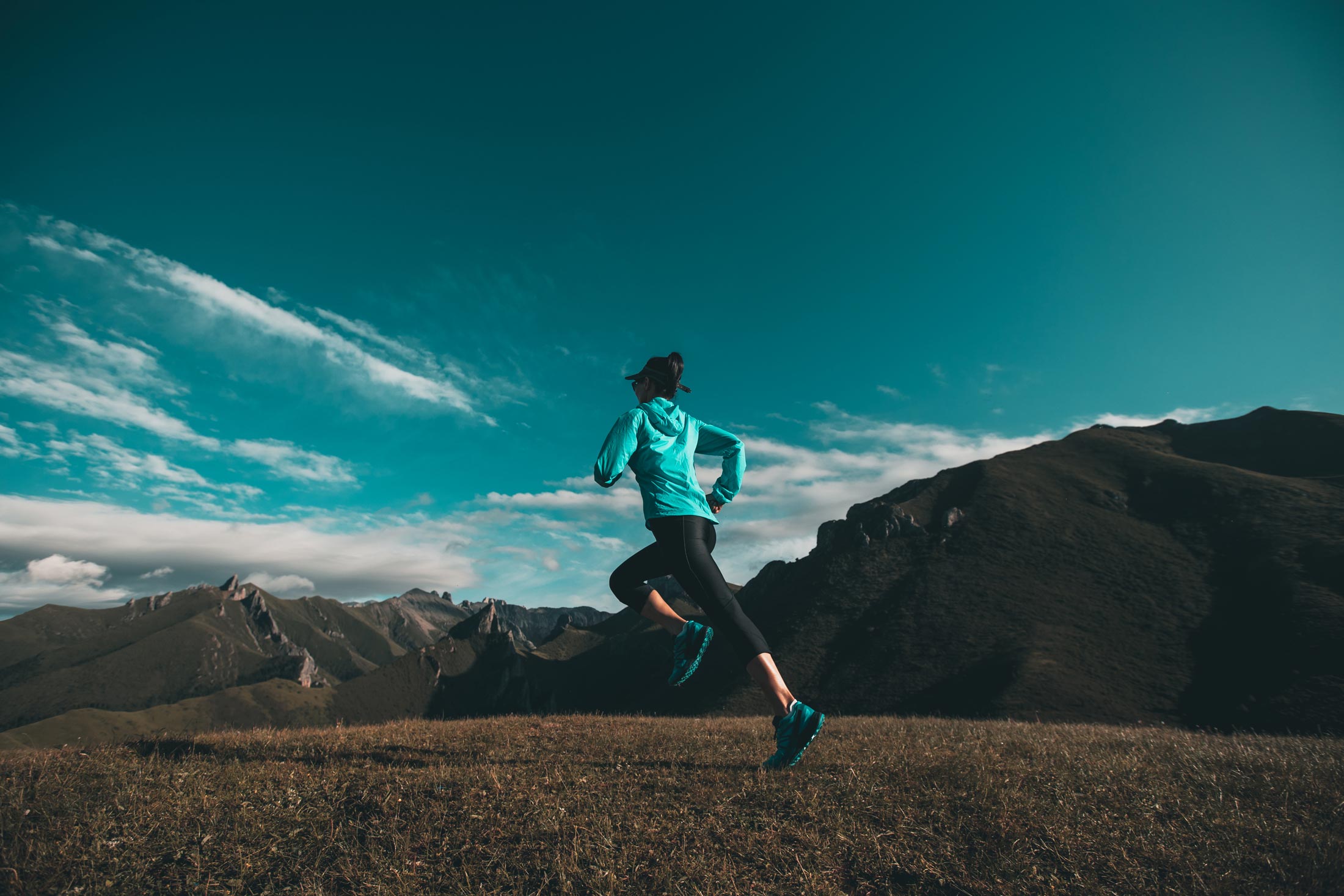 Young woman running in a turquoise jacket and black leggings on a mountain trail, emphasizing active movement and outdoor fitness.